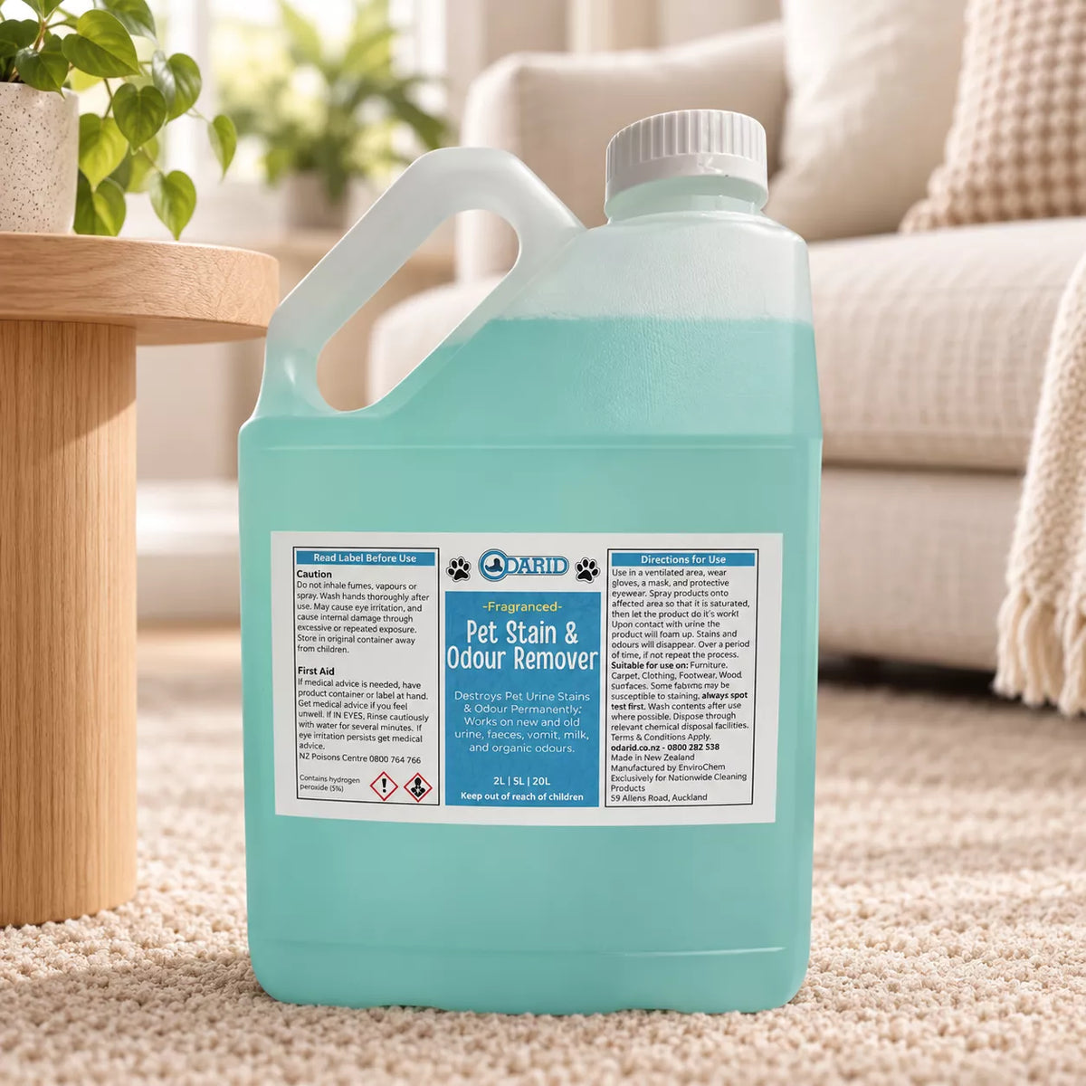 A  2 Litre bottle of Odarid Pet Stain & Odour Remover filled with light blue liquid, placed on a beige carpet in a living room setting with a sofa and side table in the background.